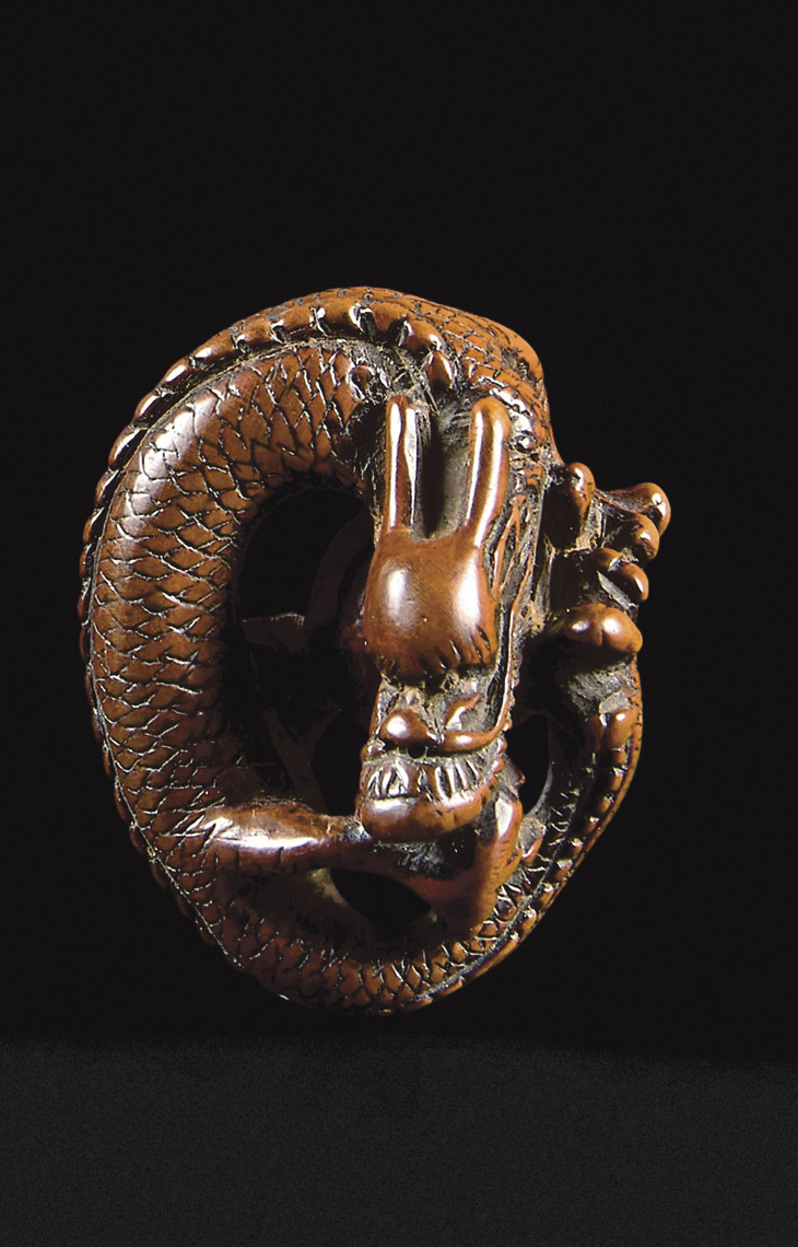 Japan, Edo period (1603-1868), 18th century. Wooden netsuke of a dragon coiled on itself, its scales finely chiseled, holding between its 