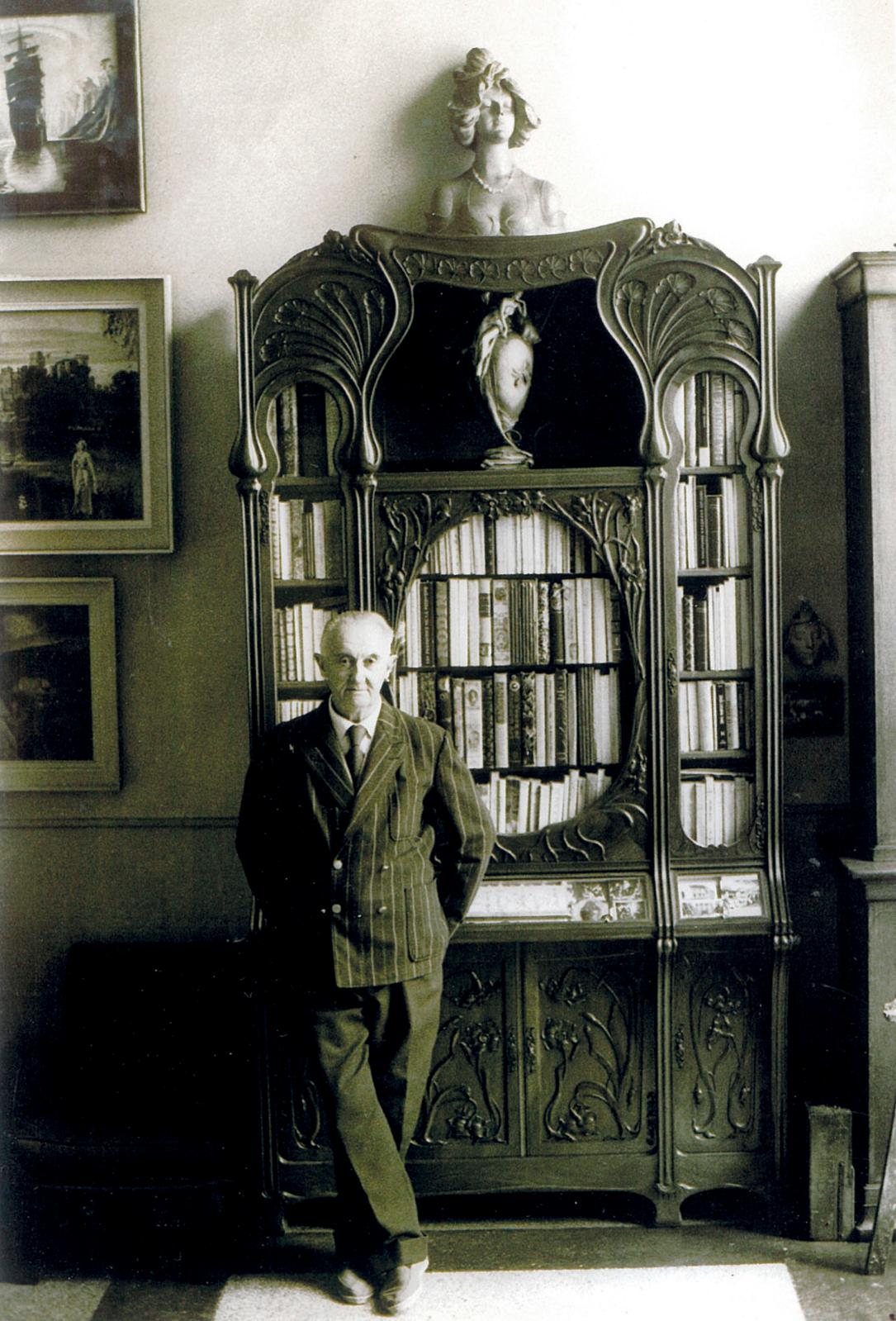 Clovis Trouille, chez lui, devant une vitrine d’exposition à deux corps en acajou, à décoren relief d’iris et d’ombelles, de l'école de Na